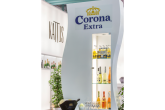 ein dreistufiges Regal mit dem Corona Logo samt kleinen und großen Corona Flaschen. im Bild unten sieht man noch einen Tisch mit Sektgläsern und einer Flasche Sekt in einem Kühlbehälter.